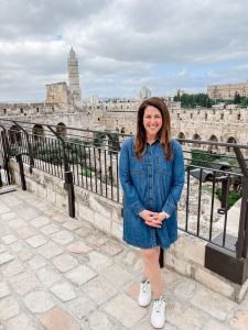 Sarah Solomon is a wearing a denim dress and standing in front on an old, white stone building.