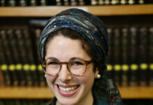 Leah Sarna is a white woman wearing a green blouse, glasses, and a blue scarf covering dark, curly hair.