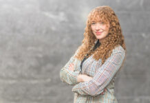 Sophia Barrett is a white woman with curly red hair. She is wearing a patterned suit jacket and posing with her arms crossed in front of a grey background.
