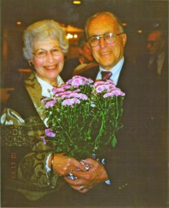 An older couple stand next to each other, smiling and holding a bouquet of flowers.